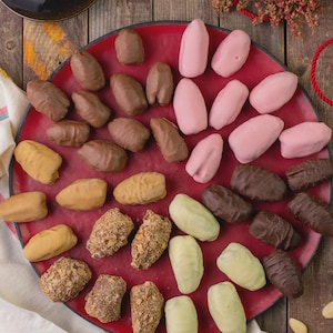 May include: A red plate holds an assortment of sweets. Some are chocolate-coated, others are covered in pink or white icing, and some are nut-covered. A cup of coffee is in the top left corner.