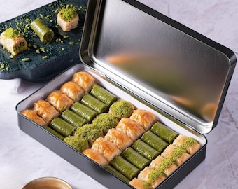 Puede incluir: Una caja metálica negra abierta llena de pasteles de baklava variados. Los pasteles son de color marrón dorado y verde, espolvoreados con pistachos. Parte del baklava está en una tabla de servir verde oscuro.