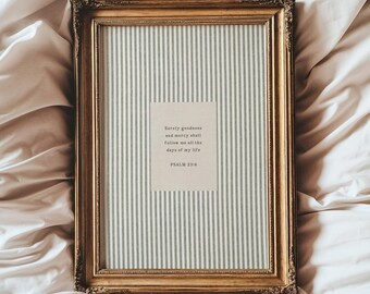 May include: A framed print with a gold-colored ornate frame. The print features a vertical striped background with a smaller rectangular section containing text from Psalm 23:6. The frame is resting on a white and beige fabric.