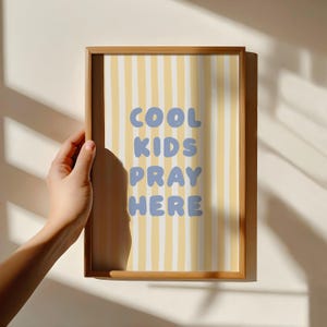 May include: A framed print with the words "COOL KIDS PRAY HERE" in blue letters. The background has vertical yellow and white stripes. The frame is made of wood.