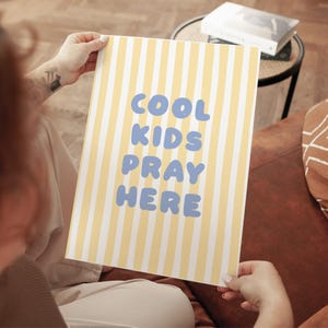May include: A rectangular poster with vertical yellow and white stripes. The poster features the text "COOL KIDS PRAY HERE" in light blue, bubble-style letters. The poster is held by a person.