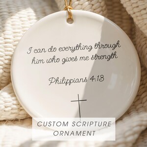 Puede incluir: Adorno de cerámica blanca con una cuerda dorada. El adorno presenta el texto "I can do everything through him who gives me strength Philippians 4:13" y una cruz. La parte inferior del adorno dice "CUSTOM SCRIPTURE ORNAMENT."
