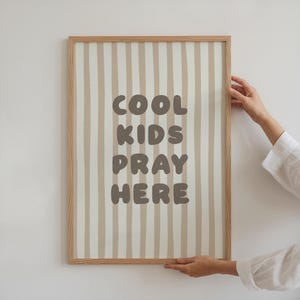 Könnte beinhalten: Ein gerahmter Druck mit einem hellen Holzrahmen. Das Kunstwerk zeigt ein vertikales Streifenmuster in Beige und Weiß. Die Worte "COOL KIDS PRAY HERE" sind in einer dunkelbraunen, abgerundeten Schrift gedruckt.