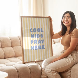 May include: A framed poster with a yellow and white striped background and the words "COOL KIDS PRAY HERE" in blue. The poster is held by a person sitting on a beige sofa.