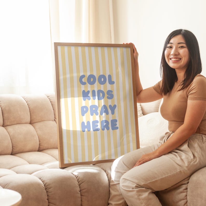 May include: A framed poster with a yellow and white striped background and the words "COOL KIDS PRAY HERE" in blue. The poster is held by a person sitting on a beige sofa.
