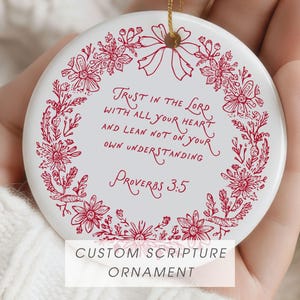 Puede incluir: Adorno redondo de cerámica blanca con diseño floral y texto en rojo. El texto dice: "Confía en el Señor con todo tu corazón y no te apoyes en tu propia comprensión Proverbios 3:5". El adorno tiene una cuerda dorada para colgar.