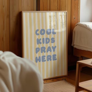 May include: Framed art print with a light wood frame. The artwork features vertical yellow and white stripes with the words "COOL KIDS PRAY HERE" in blue bubble letters. The print is displayed in a bedroom setting.