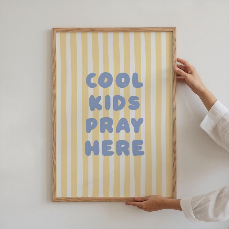 May include: A framed print with a light wood frame. The artwork features a yellow and white striped background with the words "COOL KIDS PRAY HERE" in blue bubble letters. The print is held by a person.