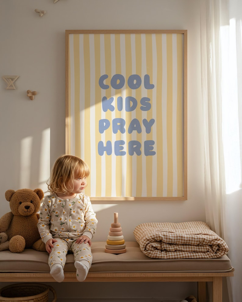 May include: A child's room with a framed poster displaying "COOL KIDS PRAY HERE" on a yellow and white striped backdrop. A young child in a floral onesie sits on a bench beside a teddy bear and a wooden stacking toy.