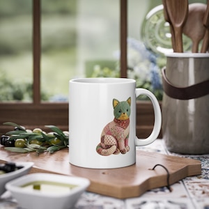 Peut inclure: Mug en céramique blanche avec un motif de chat en patchwork. Le chat a une tête à carreaux verts et bleus, une écharpe rouge et un corps fait de divers tissus à motifs. Le mug est posé sur une planche à découper en bois.
