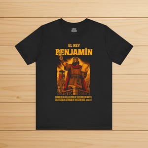 Puede incluir: Camiseta negra con un gráfico de un hombre sosteniendo un libro y gesticulando. El texto "EL REY BENJAMIN" está encima de la ilustración. Debajo, texto en español. La camiseta se muestra sobre una superficie de madera.