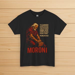 Puede incluir: Camiseta negra con un gráfico de un hombre mayor con una túnica roja, arrodillado y trabajando con una piedra. El texto "MORONI" está en rojo en la parte inferior, con texto en español arriba. La camiseta tiene cuello redondo y mangas cortas.