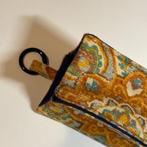 May include: A rectangular, patterned fabric item with a black ring attached. The fabric features a repeating design in shades of gold, teal, and brown. The edges are trimmed with a dark blue border. The item appears to be a small pouch or case.