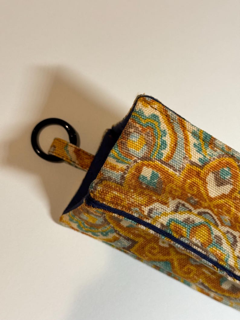 May include: A rectangular, patterned fabric item with a black ring attached. The fabric features a repeating design in shades of gold, teal, and brown. The edges are trimmed with a dark blue border. The item appears to be a small pouch or case.