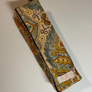 May include: A rectangular fabric case with a colorful, patterned design in shades of teal, gold, and brown. The case is trimmed with a dark blue border and features a small label with text. The case is closed with a black clasp.