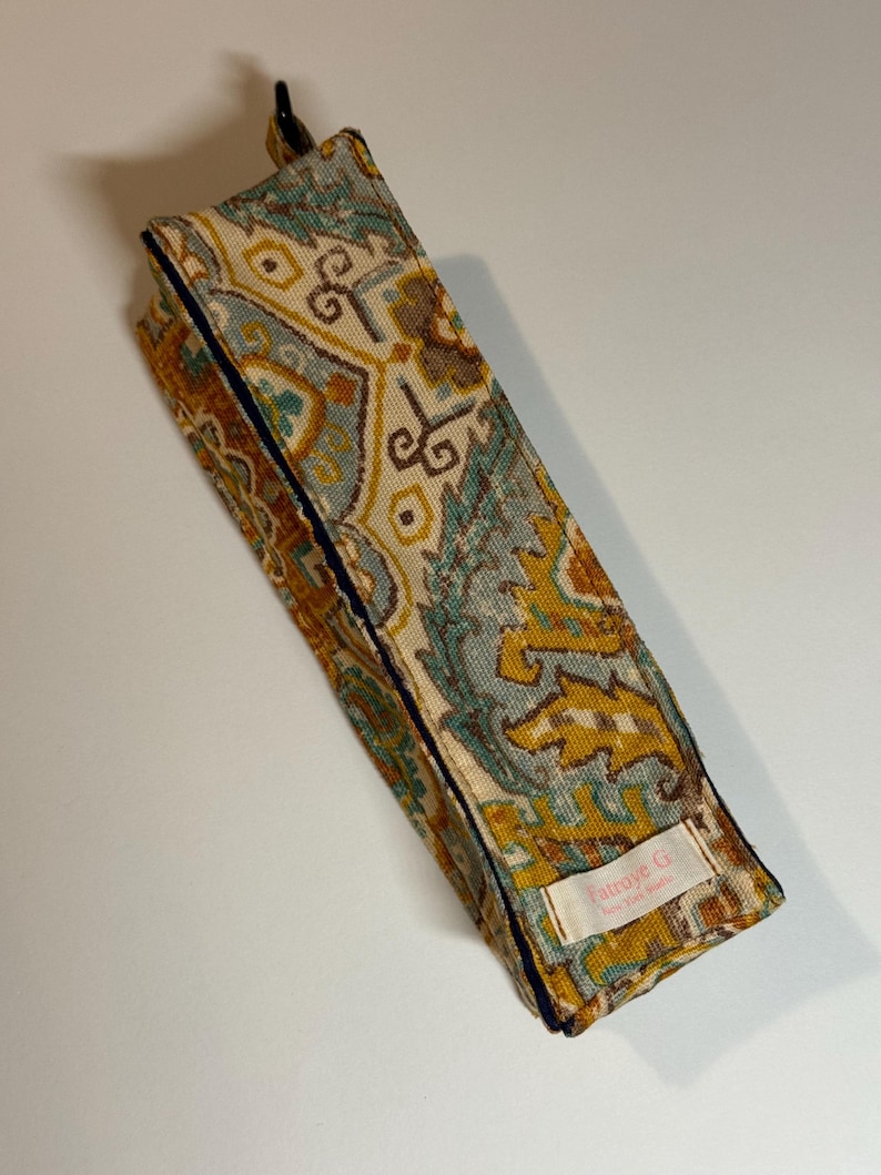 May include: A rectangular fabric case with a colorful, patterned design in shades of teal, gold, and brown. The case is trimmed with a dark blue border and features a small label with text. The case is closed with a black clasp.
