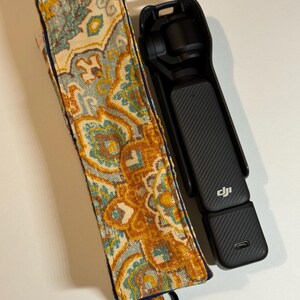 May include: A black handheld camera with a textured grip and a patterned fabric case. The case features a design in shades of yellow, teal, and blue. The camera has a USB-C port and the DJI logo.