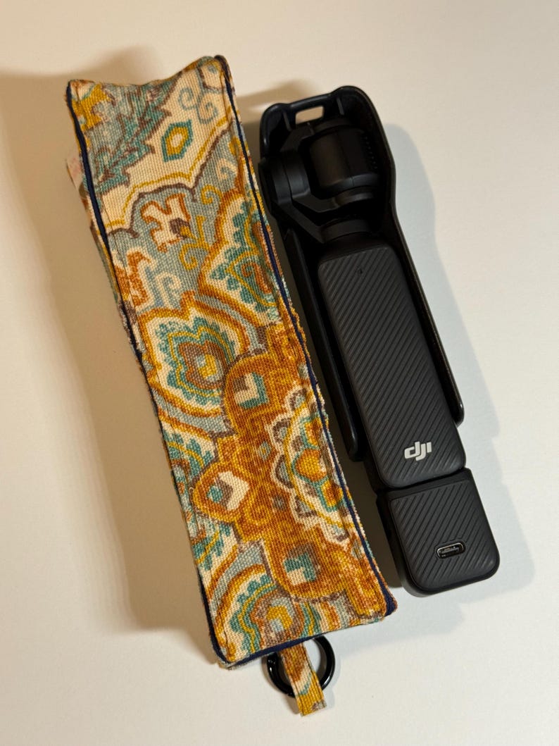 May include: A black handheld camera with a textured grip and a patterned fabric case. The case features a design in shades of yellow, teal, and blue. The camera has a USB-C port and the DJI logo.
