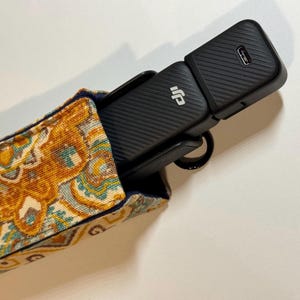 May include: A black electronic device with the DJI logo is partially inserted into a patterned fabric case. The case features a design in shades of gold, blue, and teal. The device has a ribbed texture and a USB port.