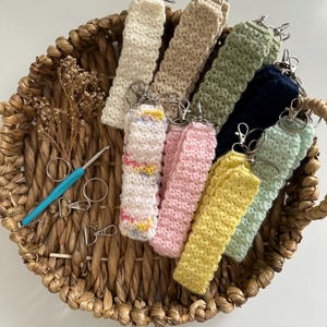 May include: A collection of handmade crochet keychains in various colors, including cream, beige, green, navy, pink, yellow, and light blue. The keychains are displayed in a woven basket with a crochet hook and scissors.