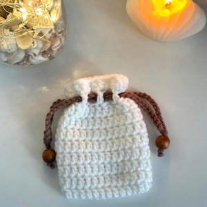 May include: A small, off-white crochet drawstring bag with brown cord and wooden beads. The bag is sitting on a white surface, with a lit seashell-shaped candle and a jar filled with seashells in the background.