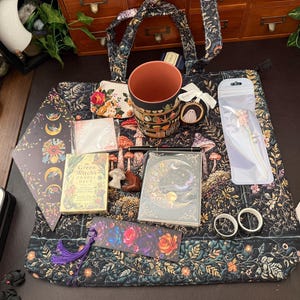 May include: A black tote bag with a floral and mushroom pattern containing a ceramic mug, a small floral coin purse, a Green Witch Oracle deck, a moon-themed notebook, a black pen, a bookmark with a purple tassel, two metal rings, and a hair accessory.