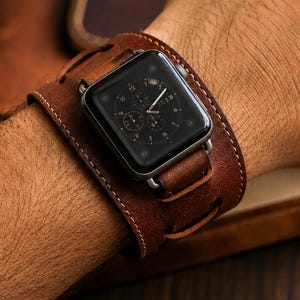 May include: A dark brown leather cuff watch band featuring a wide, wrap-around design with visible stitching and metal hardware, securely holding a rectangular digital smartwatch with a black screen displaying a circular analog clock face.
