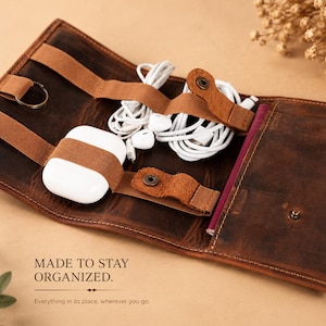 May include: A brown leather organizer case featuring elastic straps and snap-button closures to secure white wireless earbuds, white wired earphones, and a metal key ring. The interior includes a pocket and the text MADE TO STAY ORGANIZED.