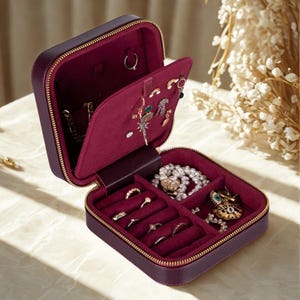 May include: A square, deep purple jewelry box with a gold zipper, featuring a velvet-lined interior in a matching dark berry shade. The case includes a hinged divider with earring holes and a base section with ring slots and open compartments.