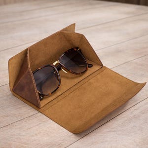 May include: A pair of brown tortoiseshell sunglasses with dark lenses resting inside an open, triangular, brown leather case. The case features a smooth, tan-colored interior lining and a structured, folding design that protects the eyewear.