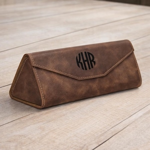 Puede incluir: Estuche de gafas de cuero marrón con forma triangular y cierre de solapa. El estuche presenta un monograma circular con las letras KHR. El estuche está hecho de cuero texturizado y tiene costuras visibles.