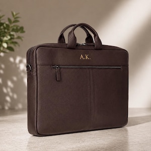 Puede incluir: Un maletín para portátil rectangular de color marrón oscuro con superficie texturizada, dos asas superiores y un bolsillo frontal horizontal con cremallera. El maletín presenta las iniciales A.K. en color dorado en la parte superior.