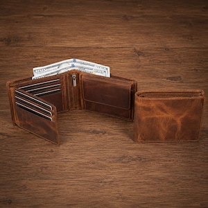 May include: A brown leather bifold wallet shown in two positions. One wallet is open, revealing multiple card slots, a zipped compartment, and US hundred-dollar bills tucked inside. The second wallet is closed, displaying a textured leather surface.