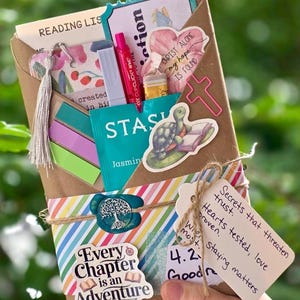 May include: A brown paper-wrapped gift package with a variety of items, including a pen, tea bag, stickers, and a tag with handwritten text. The package is decorated with colorful stripes and a twine bow. The theme is related to reading and faith.