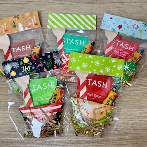 May include: Several clear cellophane bags filled with tea packets, candy, and wooden spoons. Each bag has a decorative topper with holiday-themed patterns and the text "Ho Ho Ho". The tea packets are labeled "TASH".