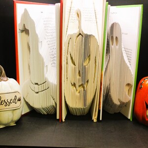 May include: Three folded books featuring paper-cut silhouettes of a witch hat, a jack-o'-lantern, and a ghost. To the left sits a white pumpkin ornament with the word blessed printed on a plaque, and to the right is an orange jack-o'-lantern.