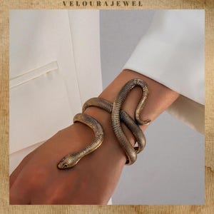 Puede incluir: Una pulsera de serpiente dorada que se enrolla alrededor de una muñeca. La pulsera presenta un diseño de serpiente detallado con escamas texturizadas y una cabeza de serpiente. La joya se usa sobre un fondo blanco.