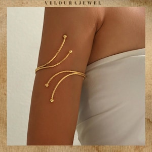 以下が含まれることがあります： ゴールドトーンのアームカフジュエリー。アームカフは、小さな球状のアクセントが付いた繊細な曲線が特徴です。ジュエリーは上腕に着用され、ニュートラルな背景に配置されています。「VELOURAJEWEL」というブランド名が上部にあります。