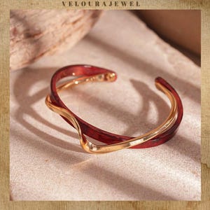 Könnte beinhalten: Ein gold-rotes Armreif-Armband. Das Armband hat ein verdrehtes Design mit einem goldenen Band, das sich durch ein rotes Band windet. Der Schmuck wird auf einer neutralen Oberfläche präsentiert. Der Text "VELOURAJEWEL" befindet sich oben.