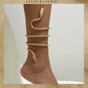 Puede incluir: Tobillera de serpiente dorada. La joya se enrolla alrededor del tobillo y la parte inferior de la pierna, con una cabeza de serpiente en la parte superior e inferior. La pulsera tiene un patrón texturizado en forma de escamas. La marca "VELOURAJEWEL" está en la parte superior.