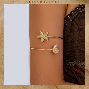 May include: Gold-toned arm cuff featuring a starfish and a seashell design. The upper cuff has a starfish, and the lower cuff has a seashell. The jewelry is worn on the arm. The background is a neutral color.