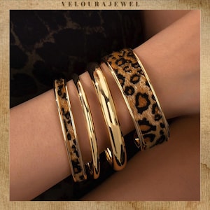 Puede incluir: Una colección de brazaletes dorados, algunos con diseño de estampado de leopardo. Los brazaletes se muestran en una muñeca, mostrando el contraste entre el oro brillante y el estampado animal texturizado. La palabra "VELOURAJEWEL" está en la parte superior.
