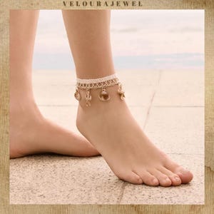May include: An off-white lace anklet featuring a woven lattice pattern and dangling gold-toned charms shaped like seashells and starfish. The delicate accessory is secured around the ankle, showcasing intricate textures and metallic accents.