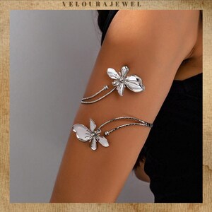 May include: Silver arm cuffs with a floral design. The cuffs feature two flower-shaped ornaments, one above the other, connected by delicate, curved bands. The jewelry is displayed on an arm, showcasing the elegant design.