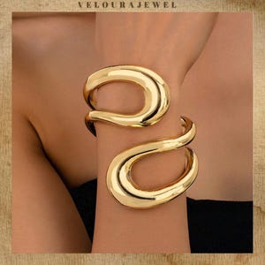 May include: A pair of gold-coloured, open cuff bracelets. The bracelets have a smooth, polished finish and a unique, flowing design. The bracelets are worn on the wrist. The brand name "VELOURAJEWEL" is at the top of the image.