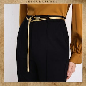 May include: A gold belt with a knot detail and a hanging chain, worn over black trousers. The image shows a mustard-coloured blouse. The brand name "VELOURAJEWEL" is at the top of the image.