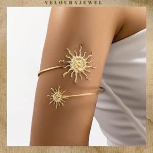 May include: A gold-toned metal arm cuff featuring two sun-shaped motifs of different sizes. The larger sun is positioned at the top and the smaller sun at the bottom, both connected by a curved, open-ended band with a textured, metallic finish.