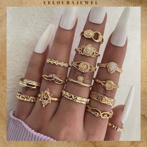 May include: A selection of gold rings on fingers with white nail polish. The rings showcase diverse designs, such as filigree, gemstones, and geometric patterns. The brand name "VELOURAJEWEL" is displayed at the top of the image.
