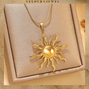 May include: Gold-coloured pendant necklace with a sunburst design. The pendant has a central circular element with radiating rays. The necklace is displayed on a neutral-toned surface, with the brand name "VELOURAJEWEL" at the top.