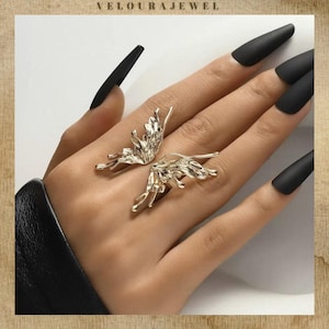 Op de afbeelding: Een goudkleurige ring met een gestructureerd, organisch vlindervleugelontwerp dat zich over twee vingers uitstrekt. Het metalen oppervlak heeft een gesmolten, onregelmatige afwerking met delicate, aderachtige details over de vleugels.